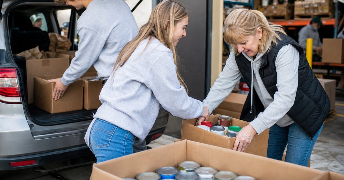 Volunteers at a food bank loading dock unpacking boxes of donated canned goods from the open trunk of a car, warehouse shelving in the background, natural daylight