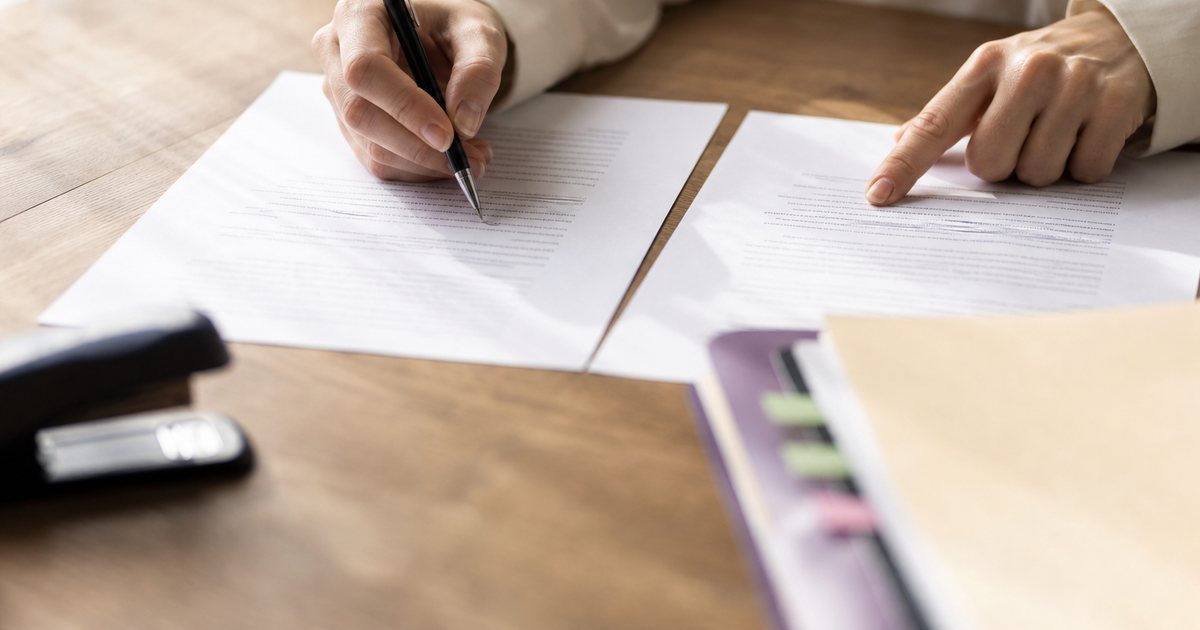Close-up of hands at a wooden desk comparing two printed letters side by side, one marked with a pen, a stapler and file folders nearby, soft window light