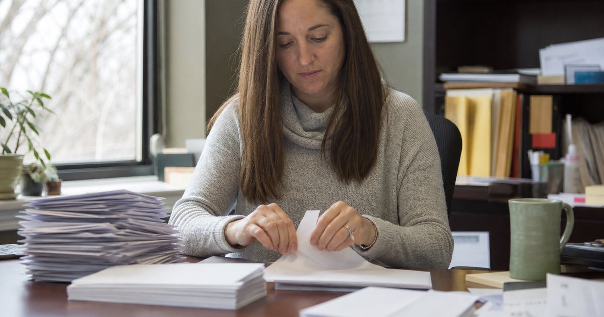 Small nonprofit office in early January, staff member sealing envelopes at a desk stacked with letters, a wall calendar and a window with bare winter trees outside